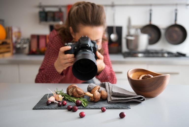 Eine Food-Fotografin im Hintergrund beugt sich vor, um in einer modernen Küche eine Nahaufnahme von Herbstfrüchten und -gemüse - Pilzen, Knoblauch, Rosmarin und Preiselbeeren - zu machen.