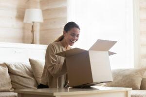 Lachende junge Frau schaut in einen offenen Karton, packt das Paket aus, sitzt auf der Couch im Wohnzimmer, zufrieden und aufgeregt über das Geschenk