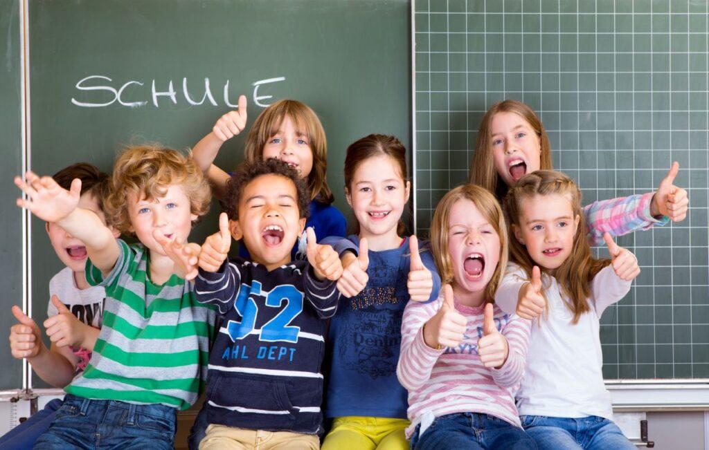 Gruppe lachender Kinder vor einer Tafel in einer bilingualen grundschule berlin mit dem Schriftzug „Schule“ im Hintergrund